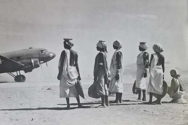 Black and white photograph of a group of six Manipuri women standing at a distance from military aircraft on a dusty airfield. They are holding baskets, and have their hair tied in wraps. Some have caps that enable them to carry loads on their heads.