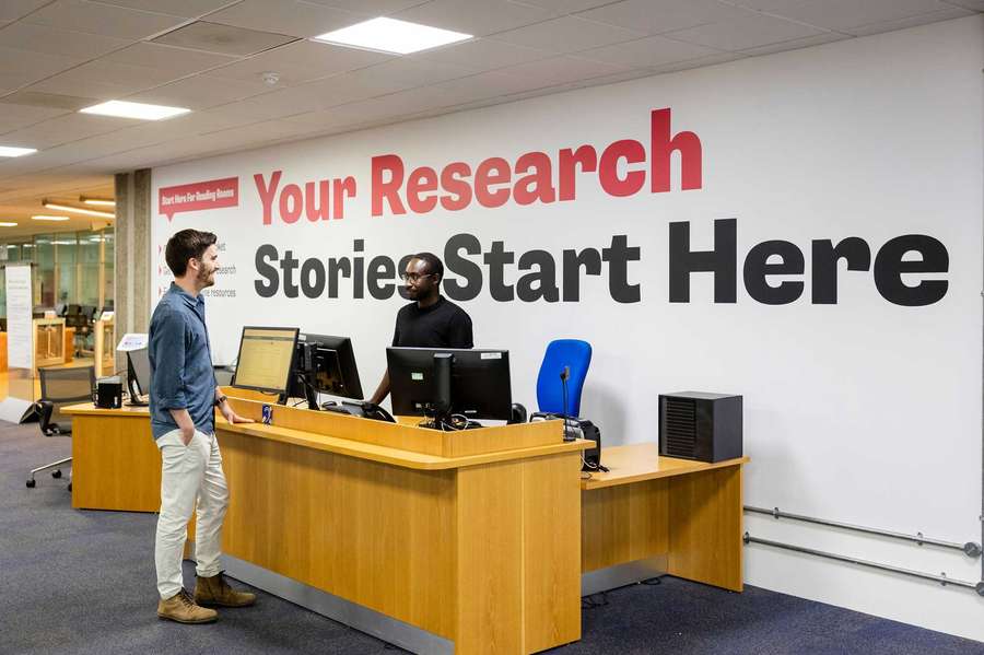 A visitor is helped by a member of staff at a desk with a large sign behind it saying 'Your research stories start here'.