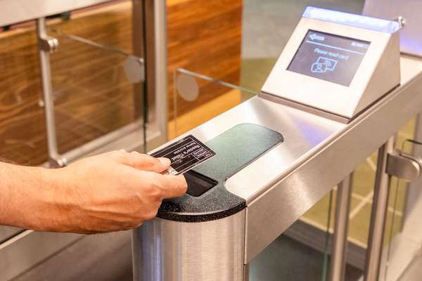 A visitor's hand holds a small, black plastic card over a scanner in front of a closed gate.