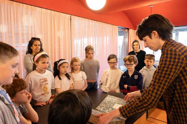 A class of children gather round a large book lying open on foam supports, held by a member of staff.
