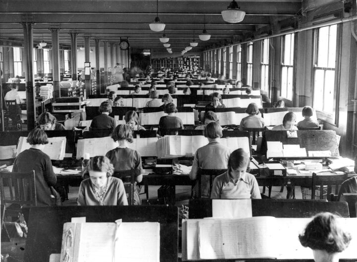 Interior of a large office with windows on each side filled with a least ten rows of desks. Women sit either side of each long desk working on large volumes containing the census schedules.