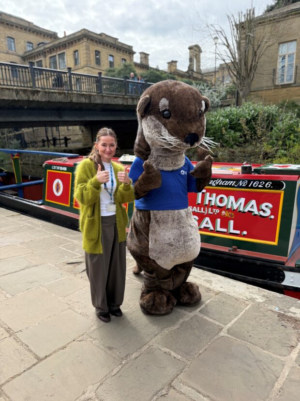 A woman and a person in a large otter mascot costume stand next to each other giving a double thumbs up, on a canal path with a canal boat and bridge in the background.