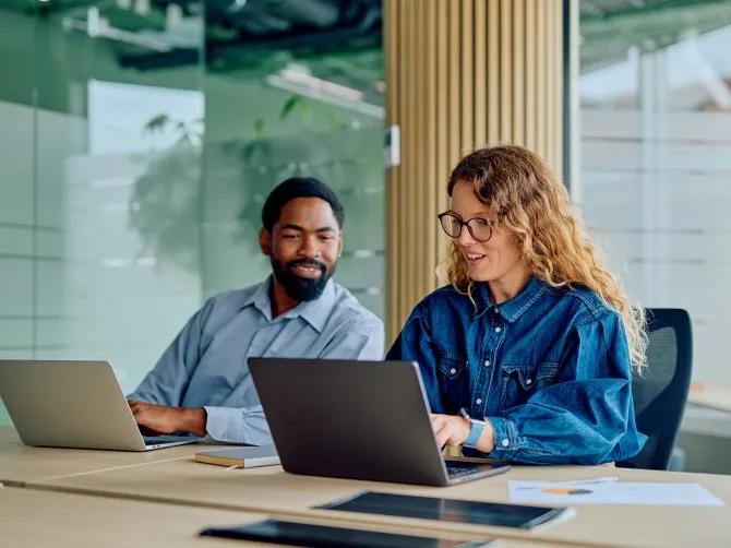 A smiling man and woman sit at a desk. They look relaxed. The woman is typing something into a laptop.