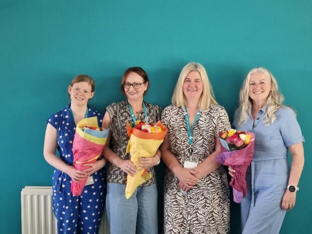 Four women in smart casual clothing and blue lanyards stand against a blue wall, smiling and holding bouquets of flowers.