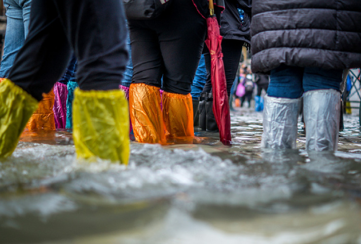People wearing brightly coloured waterproof boot covers wade through floodwater on a flooded street. Water reaches above ankle height, and a red umbrella is visible among the crowd.