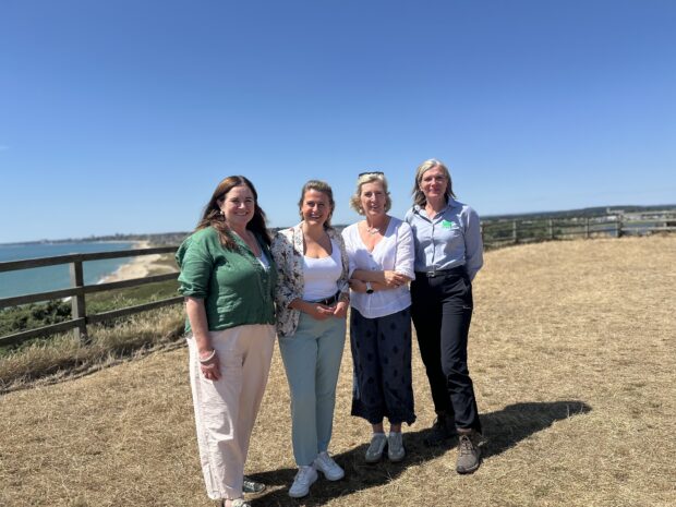 Photo showing 4 smiling women in smart casual clothing standing in a row looking at the camera, in a field with the coastline in the background.