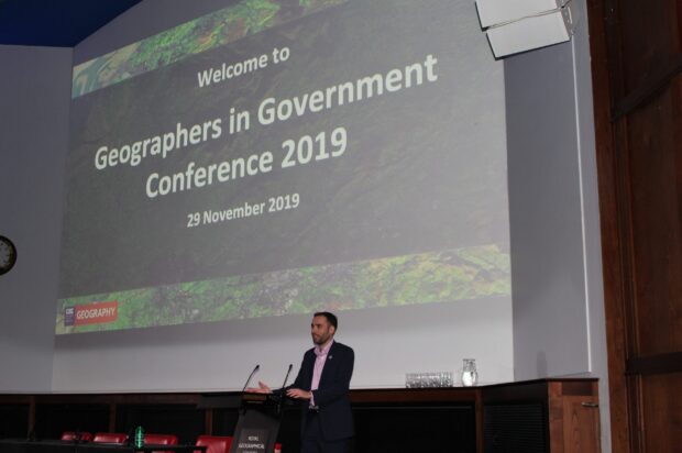 Man on stage talking in a mic. Text on screen says "Welcome to Geographers in Government Conference 2019 29th November 2019". With a Geography logo showing in the bottom left of the screen.
