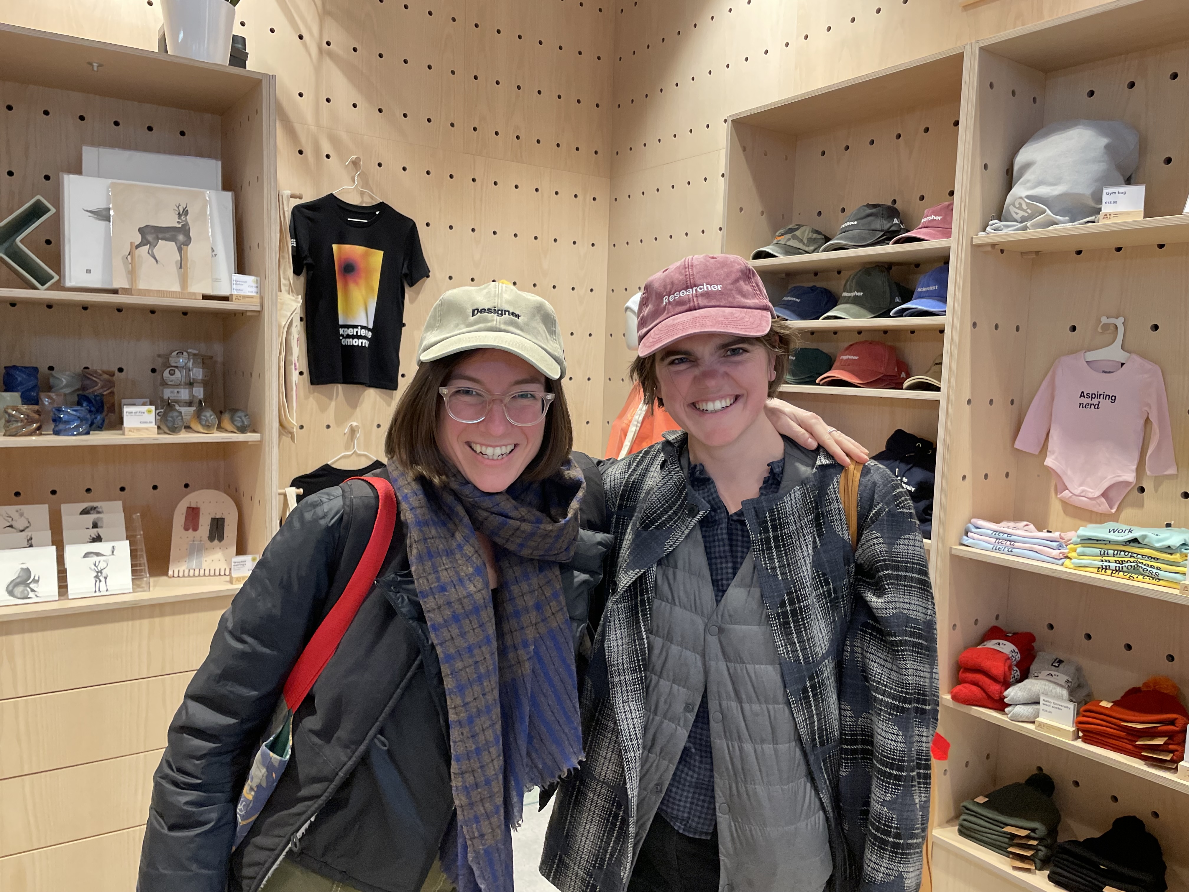 Juliette and Sarah at the International Design in Government Conference in Helsinki. They are posing smiling together. Juliette is wearing a ballcap that says 'designer' and Sarah is wearing the same style that says 'researcher'