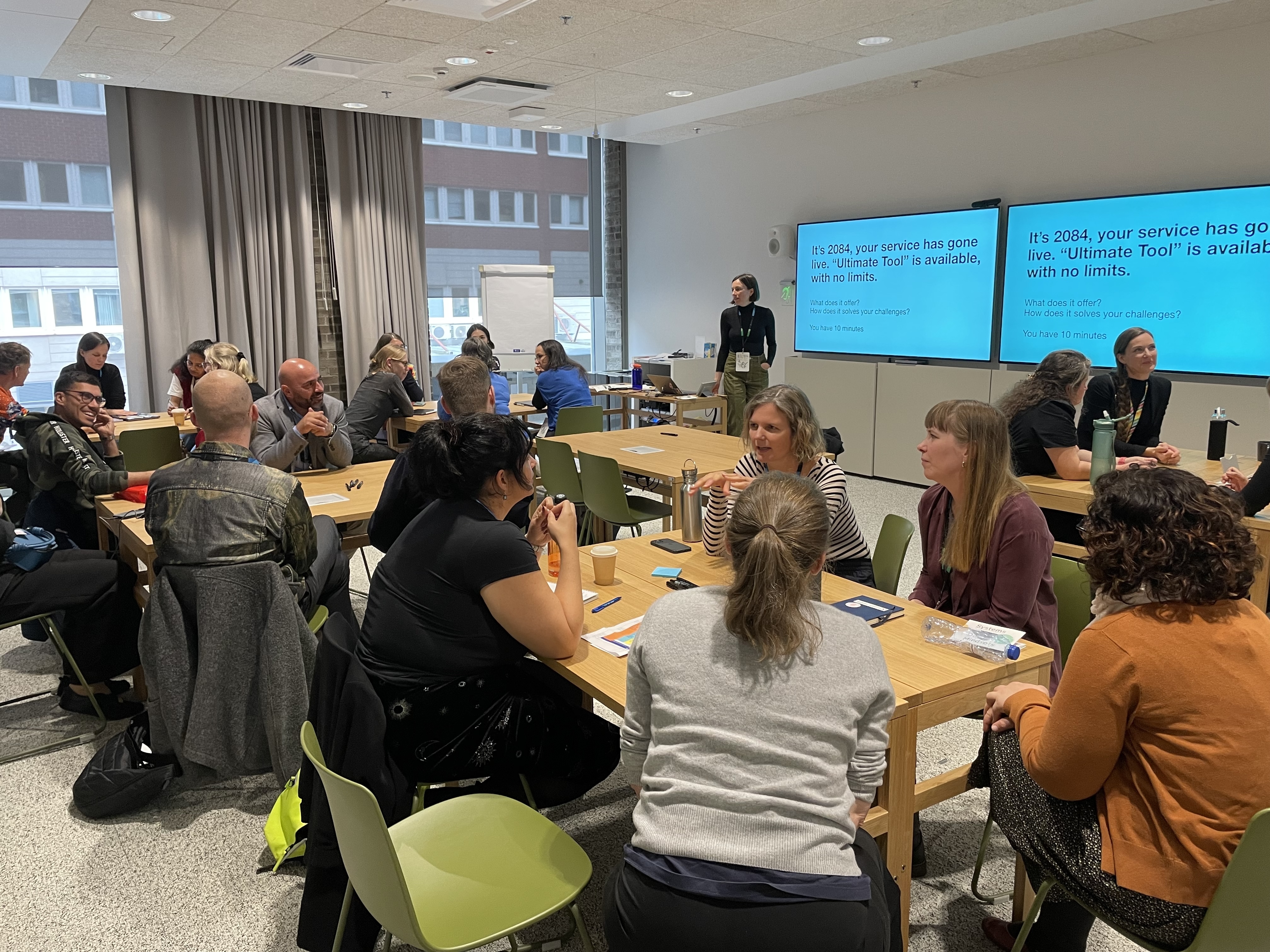 Conference attendees in a room sitting around tables in groups of 4 or 5. People are engaged in discussion in their groups. At the front of the room is one of the presenters standing in front of a screen watching on as people discuss.