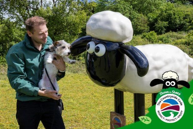 Image shows Tim and Roo from Stokenchurch Dog Rescue with The Countryside Code Champion, Shaun the Sheep statue. Tim hold Roo in his arms whilst they look at Shaun in a grassy field.