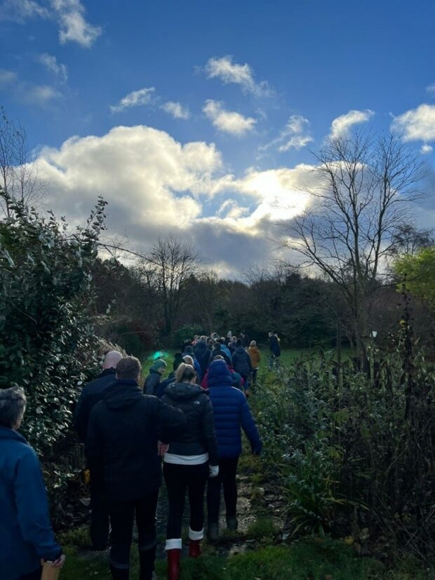Image shows a small group of young people engaging in nature activities. They are walking single file along a muddy track through an opening in the trees - likely a park or country walk. A blue sky hangs overhead with a large fluffy cloud that has caught the sun on one side, it shines bright white - the young people are all wearing coats, indicating that it's cold outside and late autumn.