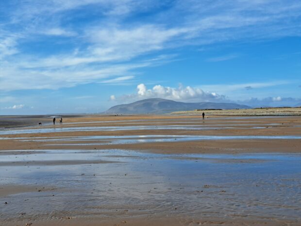 Image is taken from the beach, the tide is out and so a vast open sandy shore stretches for miles. Patches of seawater still linger, reflecting the bright blue sky and clouds above. In the far distance is a small mountain, a fluffy cloud circles it. A few people are dotted along the shore as they take a stroll.