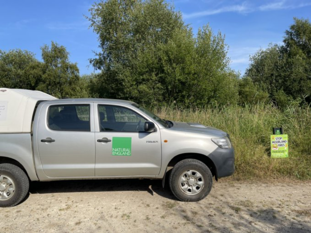 A silver pick up truck is parked at the nature reserve. The truck has a green Natural England logo on the side.