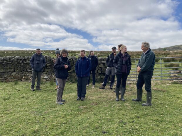 A group of people in wellies stood in a field next to a dry stone wall and gate, chatting and laughing. One is smiling directly at the camera.
