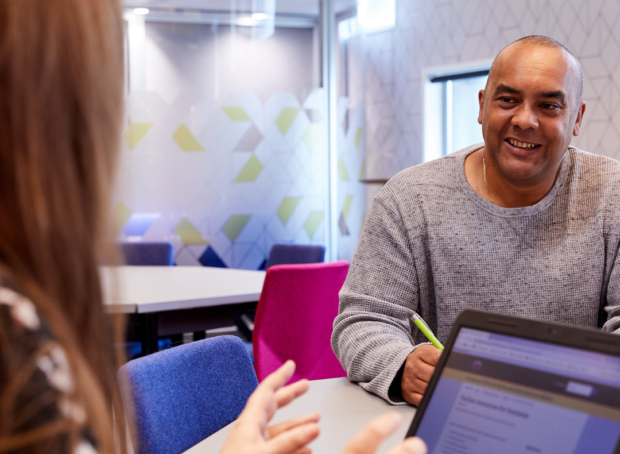 An image of two people having a conversation over a table, one with a laptop open.