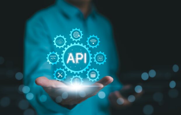 A person holds out their hand with a glowing graphic above it showing the word ‘API’ surrounded by blue gear icons representing technology concepts such as coding, security, networking and data.