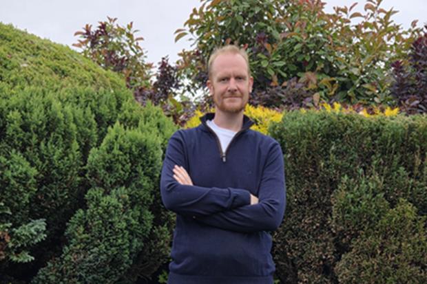 A man with a beard and short fair hair, standing, wearing a blue fleece, with arms crossed, and bushes in the background.