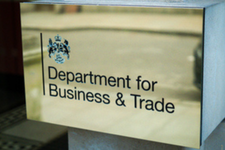 Metal plaque mounted on a building wall displaying the UK government crest and the text “Department for Business & Trade” in black lettering on a brushed metallic background.