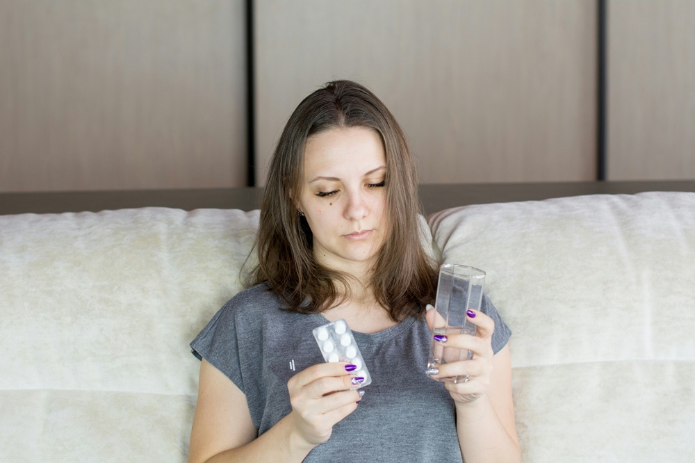 Woman on a sofa holds a packet of pills in one hand and a glass of water in the other. (Credit: Bermix Studio, Unsplash)