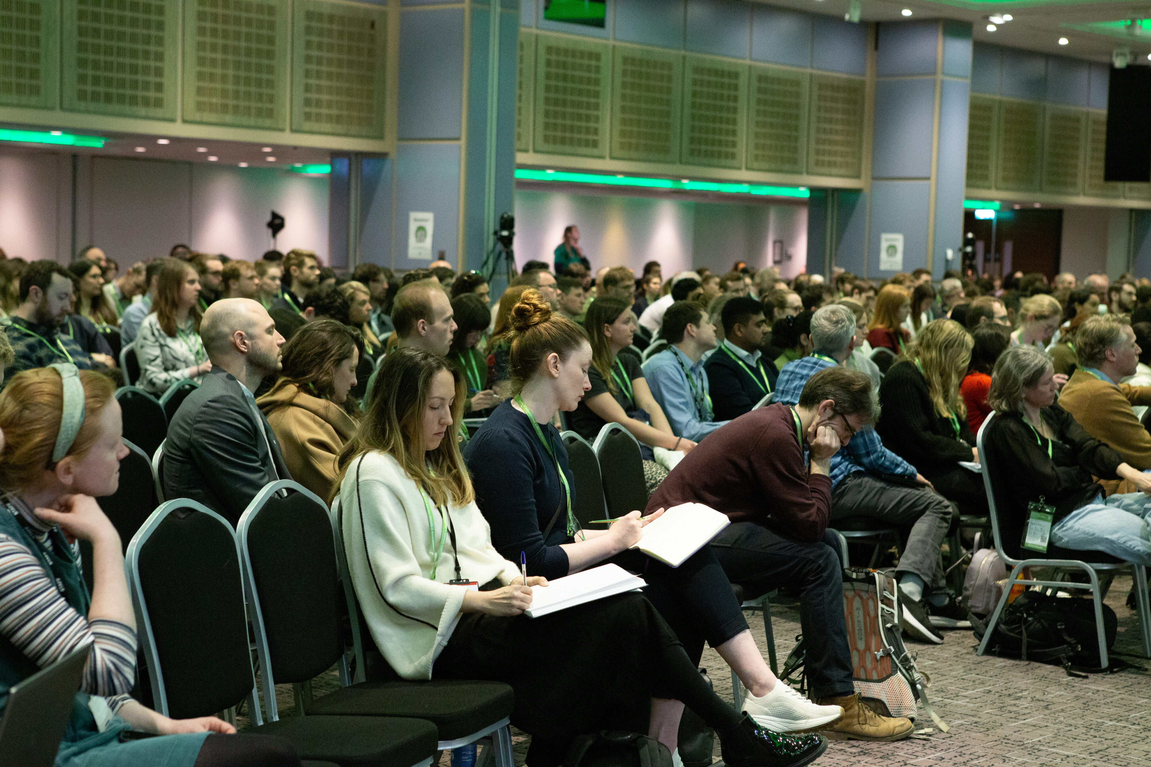 View of a conference hall where hundreds of people are attending an event. The hall is lit by green light.