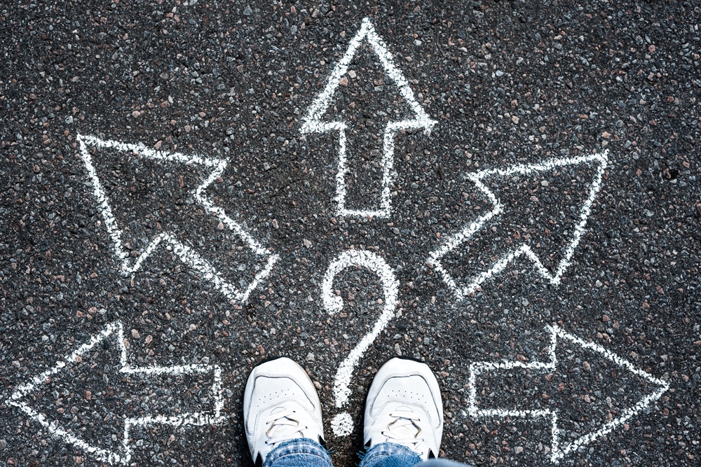 View looking down to road surface from person wearing white trainers. In front of the shoes is a large question mark, with 5 arrows branching out from there. Credit: Shutterstock