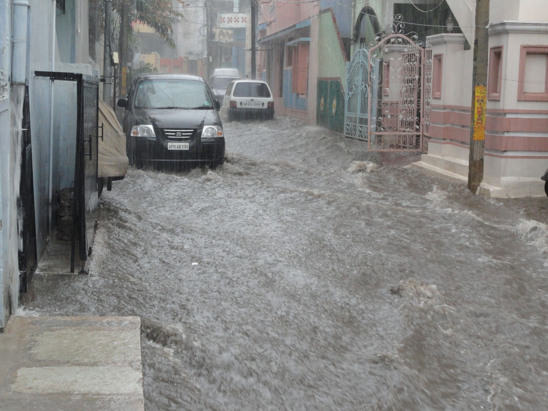 Floodwater rushing down the centre of a town centre road, Cars are parked either side of the road and it is still raining.