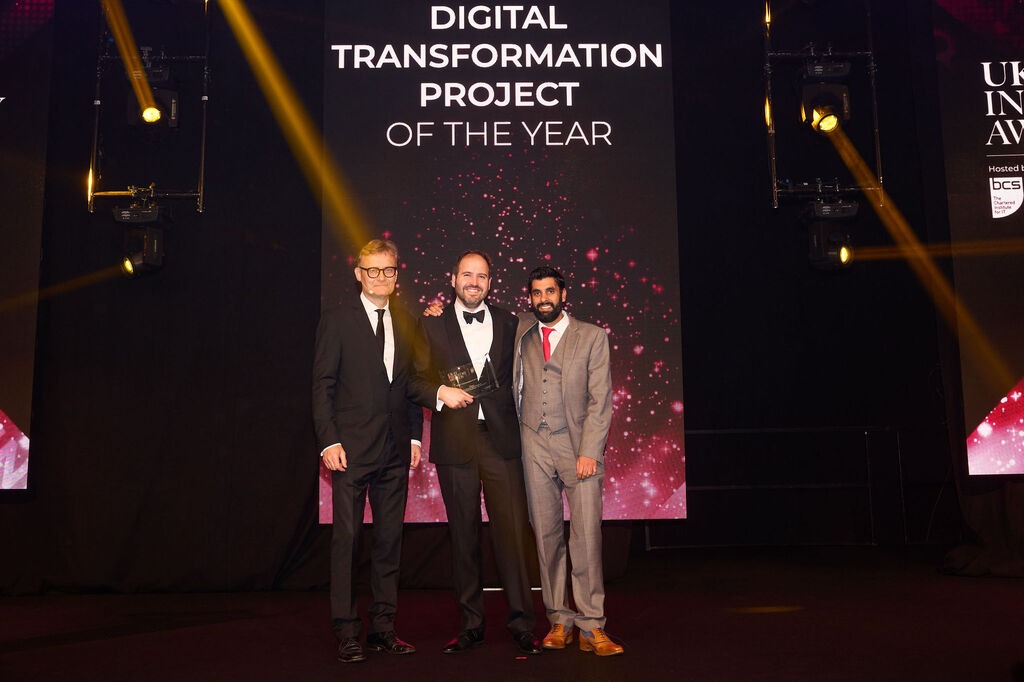 Three men in formal attire stand on stage, with one holding an award, in front of a screen that reads "Transformation Project of the Year."