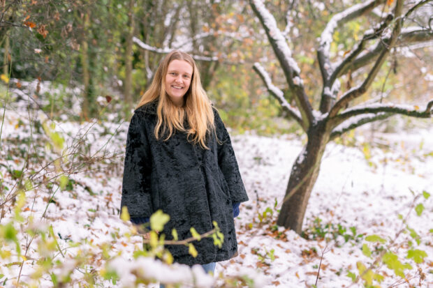 Image shows Ecologist Apprentice, Annalise Machin facing the camera, she is stood in a snow covered forest. A tree stands behind her. She wears a thick, black furry coat.