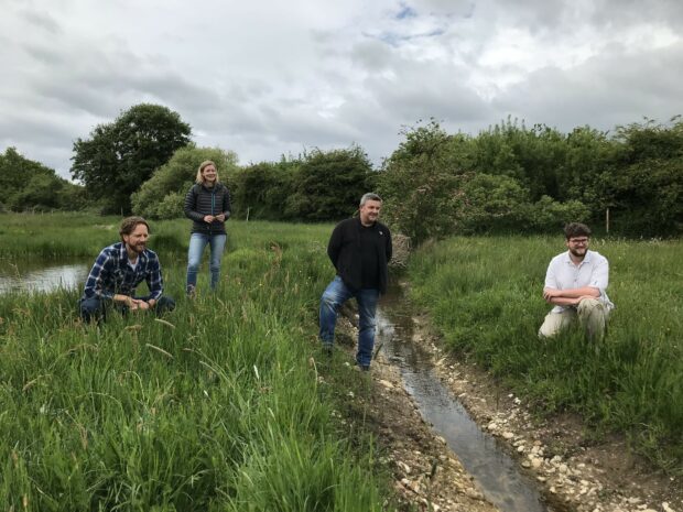 Four staff members stand and crouch at the side of the pond, next to a small stream. They are surrounded by grasses, and trees stand in the background. A grey sky hangs overhead.