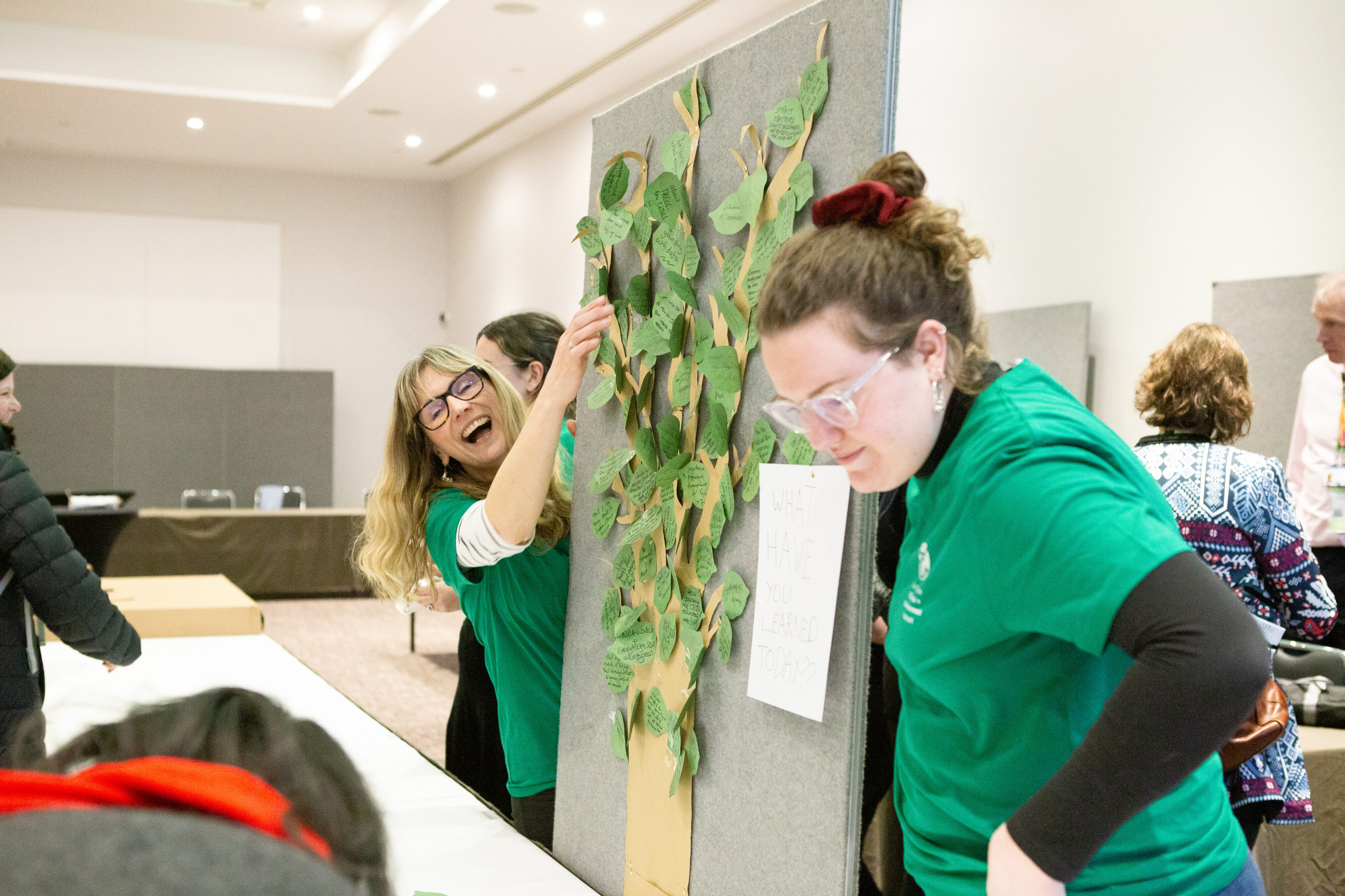 Two women wearing green t-shirts are fixing paper leaves to a paper tree which has been pinned onto a large grey hessian board.
