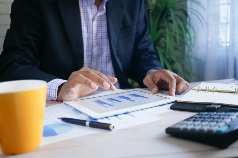 Businessman sitting at a desk looking at bar charts on a tablet. In front of him is a small ring binder file, a calculator, a pen and a yellow mug.