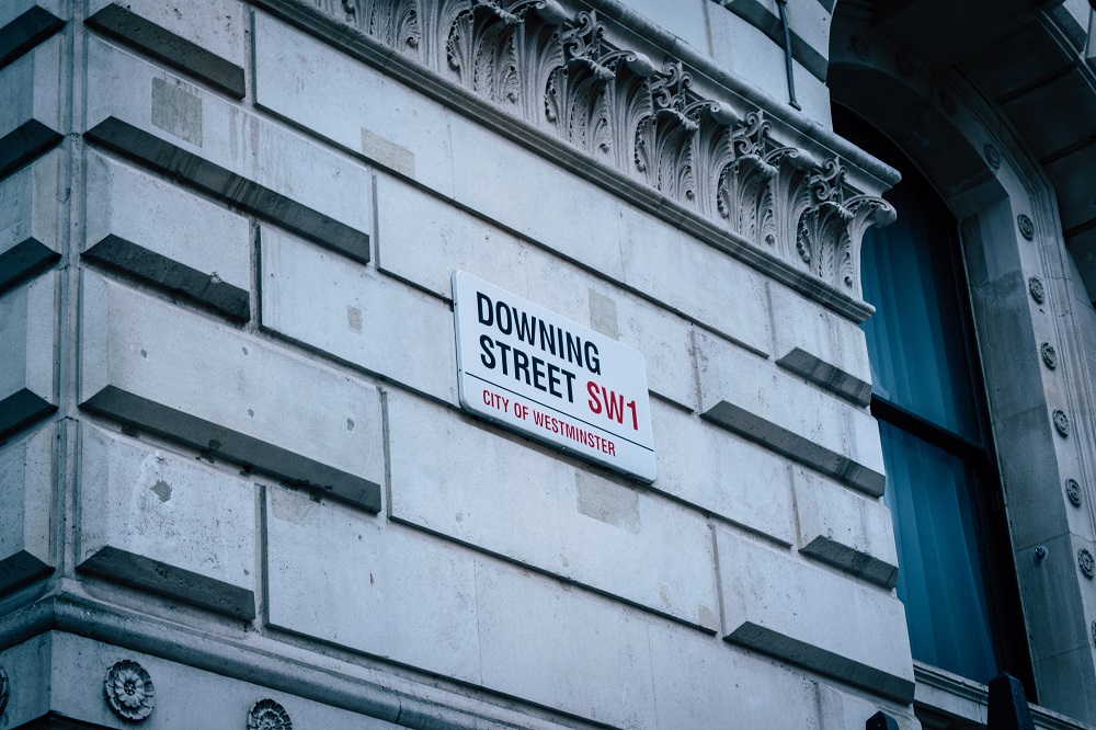 The road sign 'Downing Street SW1' is pictured on an off white exterior wall. Above the sign there is ornate cornicing and there is a window to the right of the image.