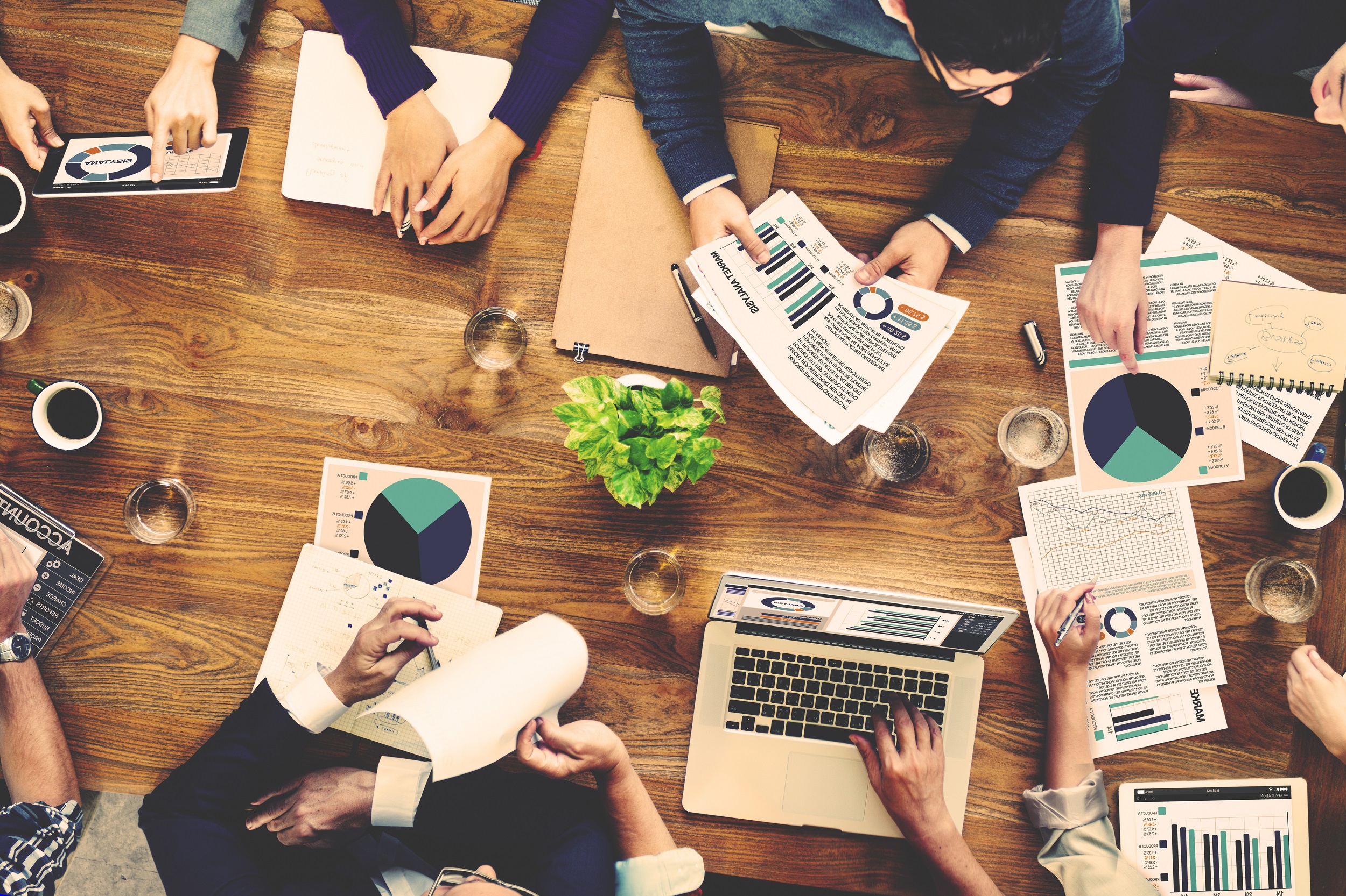 Image shows top view of a meeting with a group of 5 people using laptops, papers and notes at a wooden table.