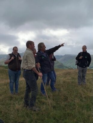 Tony stands with a group of sheep farmers to learn more about the Cumbria Common. The image is dark and grey, with deep grey clouds looming overhead, warning of possible rain. Tony points at something in the far distance.