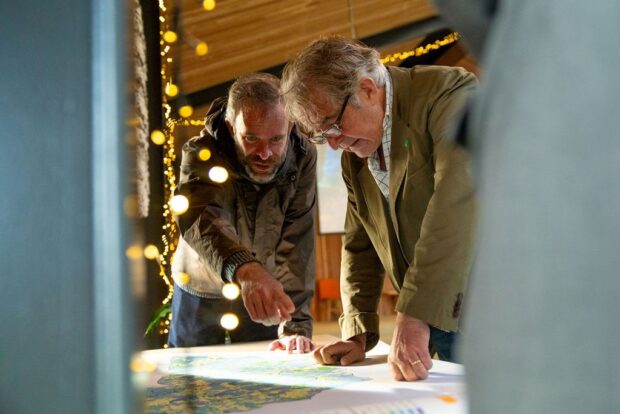 Tony looks over a map with a project lead inside a farm building. A string of warm yellow fairy lights hangs in the corner of the frame of the photo.