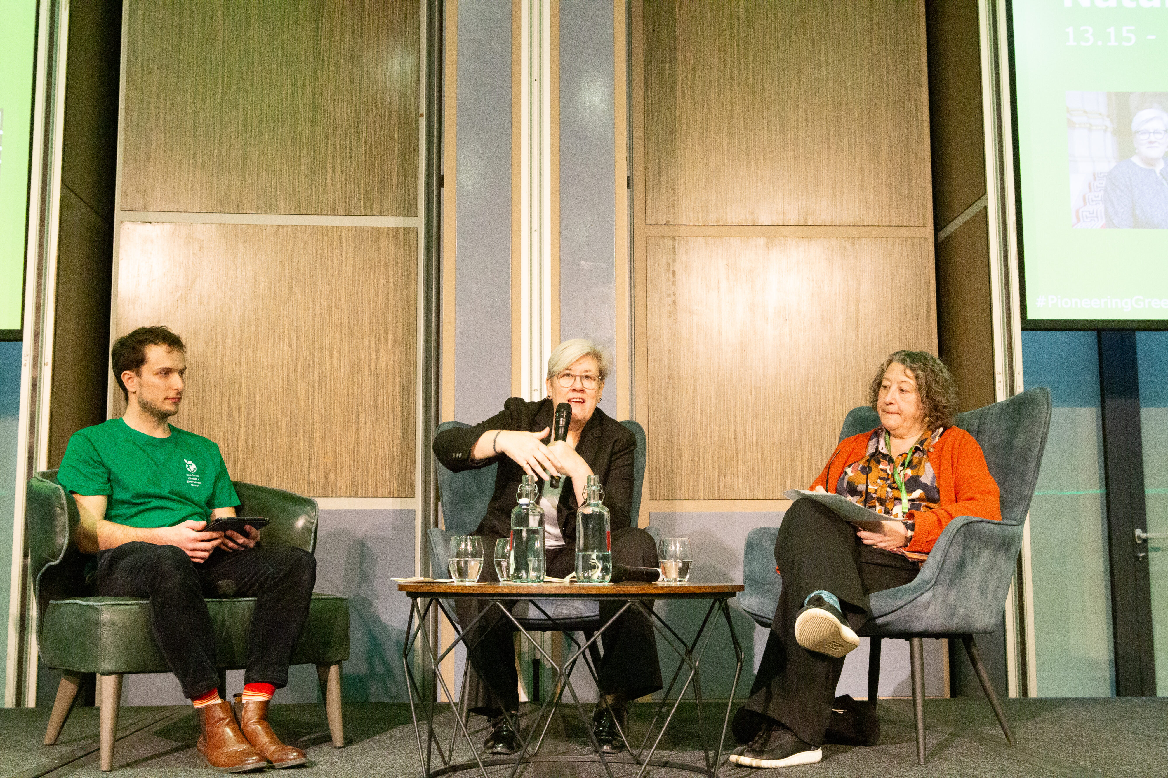 Three people (a man and 2 women) sit on a dais with a small table in front of them, on which there are bottles of water.