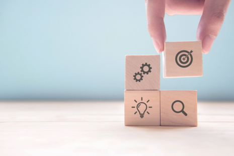 A hand arranging a wood block with a target icon on it, on top of three other wood blocks which have icons of gears, a lightbulb and a magnifying glass.