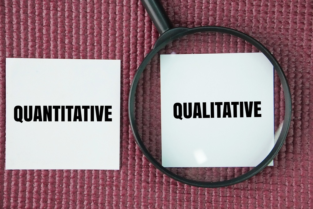 Magnifying glass on a pick textile background. It is placed above a white square with the word 'Qualitative' on it. Next to the magnifying glass is a white square with the word 'Quantitative'. Credit: iStock Photo.