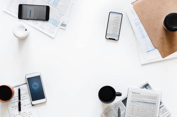 Image shows a table which has some phones,cups filled with liquid, some papers and a pen.