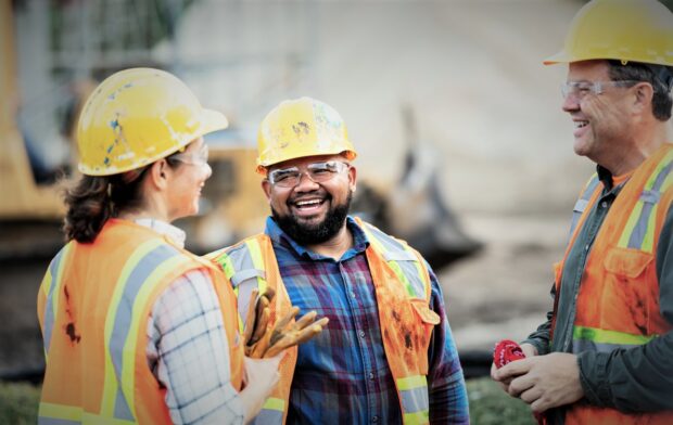 A group of three workers at a construction site wearing hard hats, safety glasses and reflective clothing, smiling and conversing.
