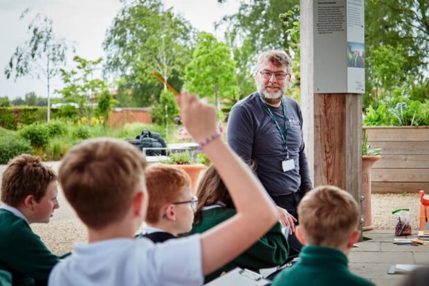 Children stand in front of a teacher, as they study about nature. One child has his hand raised. There are vivid trees and greenery in the park behind them.