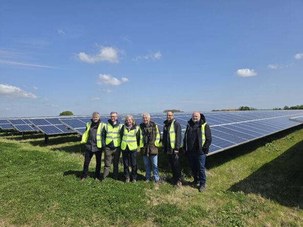 Six people in outdoor clothing and high vis jackets pose for a photograph in front of a field of large solar panels.