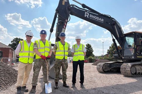 A group of four people in high vis jackets and helmets stand in front of a digger.