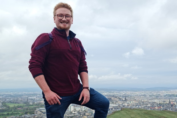 Photograph of a person standing on a hill overlooking a cityscape under a cloudy sky. The individual wears a maroon pullover and blue jeans, with one leg raised on a rock, capturing a scenic outdoor moment.