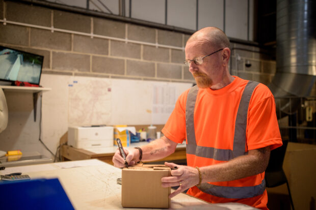A male worker wearing a hi-vis t-shirt. He is stood at a worktop labelling a box.,