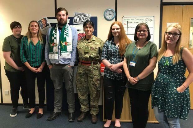 7 reservist staff members stand in a row, the middle person wearing a camouflage uniform, while the others all have green items of clothing on.