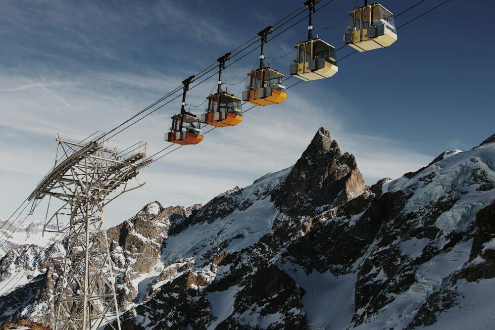 A line of 5 cable cars all in a row, pictured against a rocky mountain background, some of it covered in snow. There is a blue sky, with some high cirrus clouds.