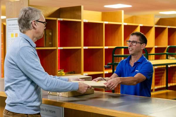 A visitor holds two boxes of records and a volume to a member of staff at the Document Reading Room returns desk.