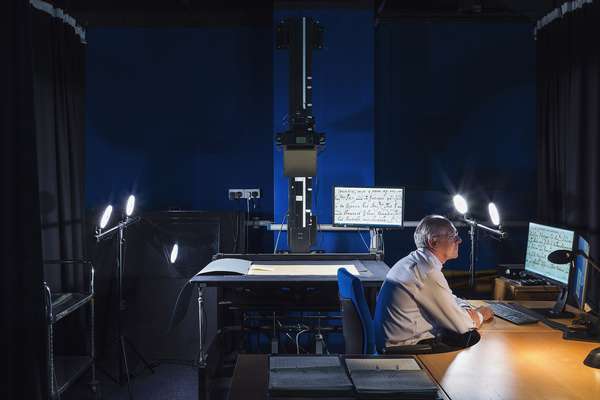 A worker sits at a desk in a dark room looking at a photograph of a historical document on a screen. Next to him on a table under a camera is the document.