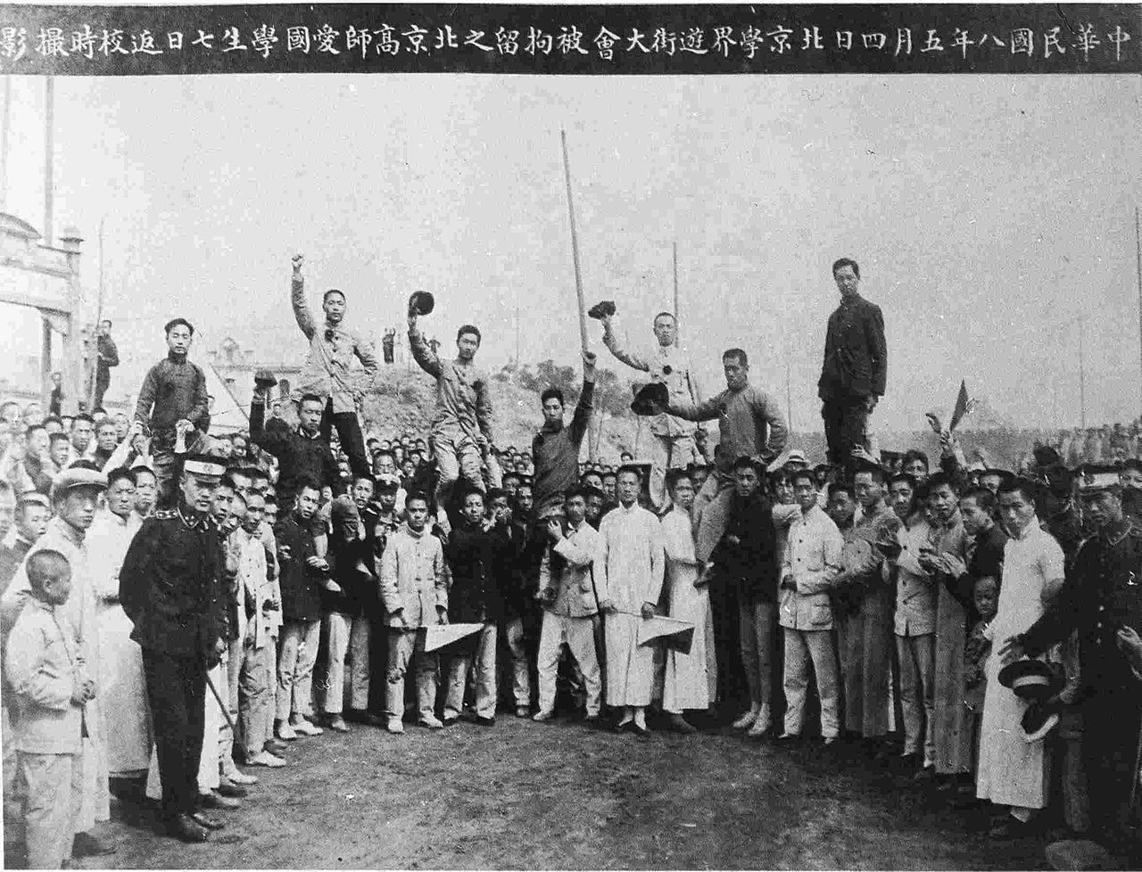 A group of university students in Beijing pose together in a big group. Seven students stand or sit on other students' shoulders and most of them raise their fists or hats in the air. Two uniformed guards are seen standing in front of the crowd, one holding out his hand as if he is holding them back.